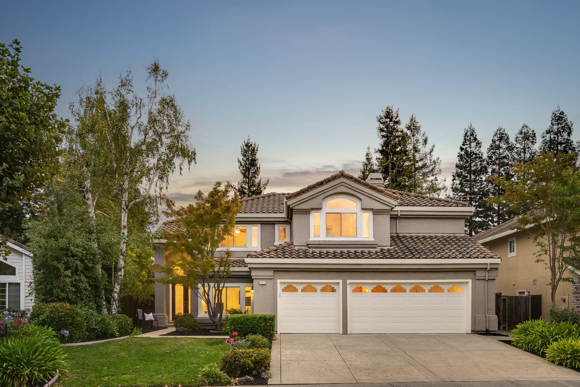 The two-story home at dusk with lit windows, a three-car garage, and a landscaped front yard.